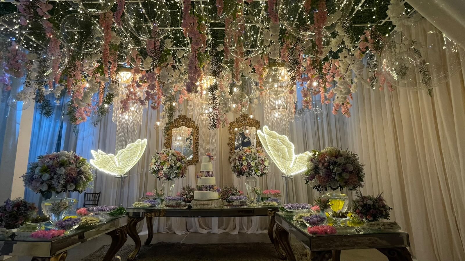 Teto de flores, lustres de cristal e borboletas iluminadas sobre a mesa de doces no Espaço Boulevard